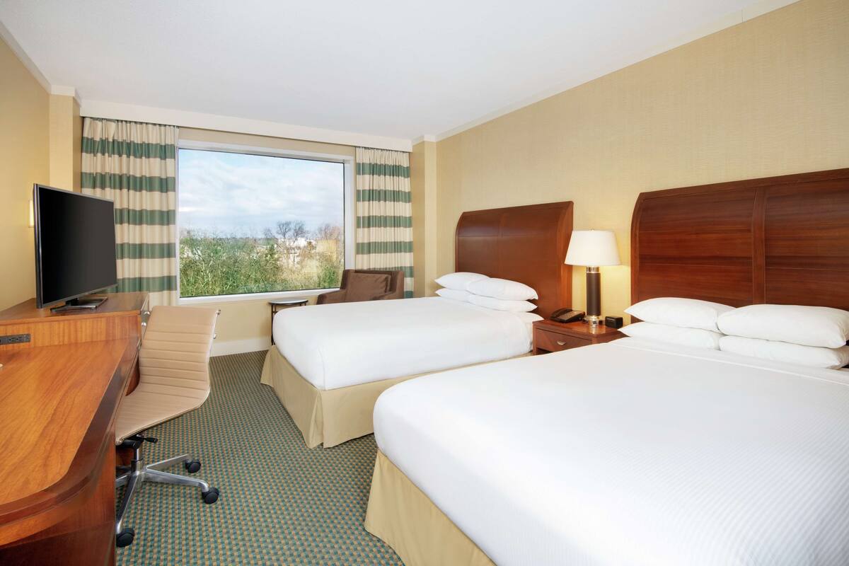 Ein Hotelzimmer mit zwei Betten, einem Schreibtisch mit Fernseher, einem Sessel, einer Lampe und einem Fenster mit Vorhängen.