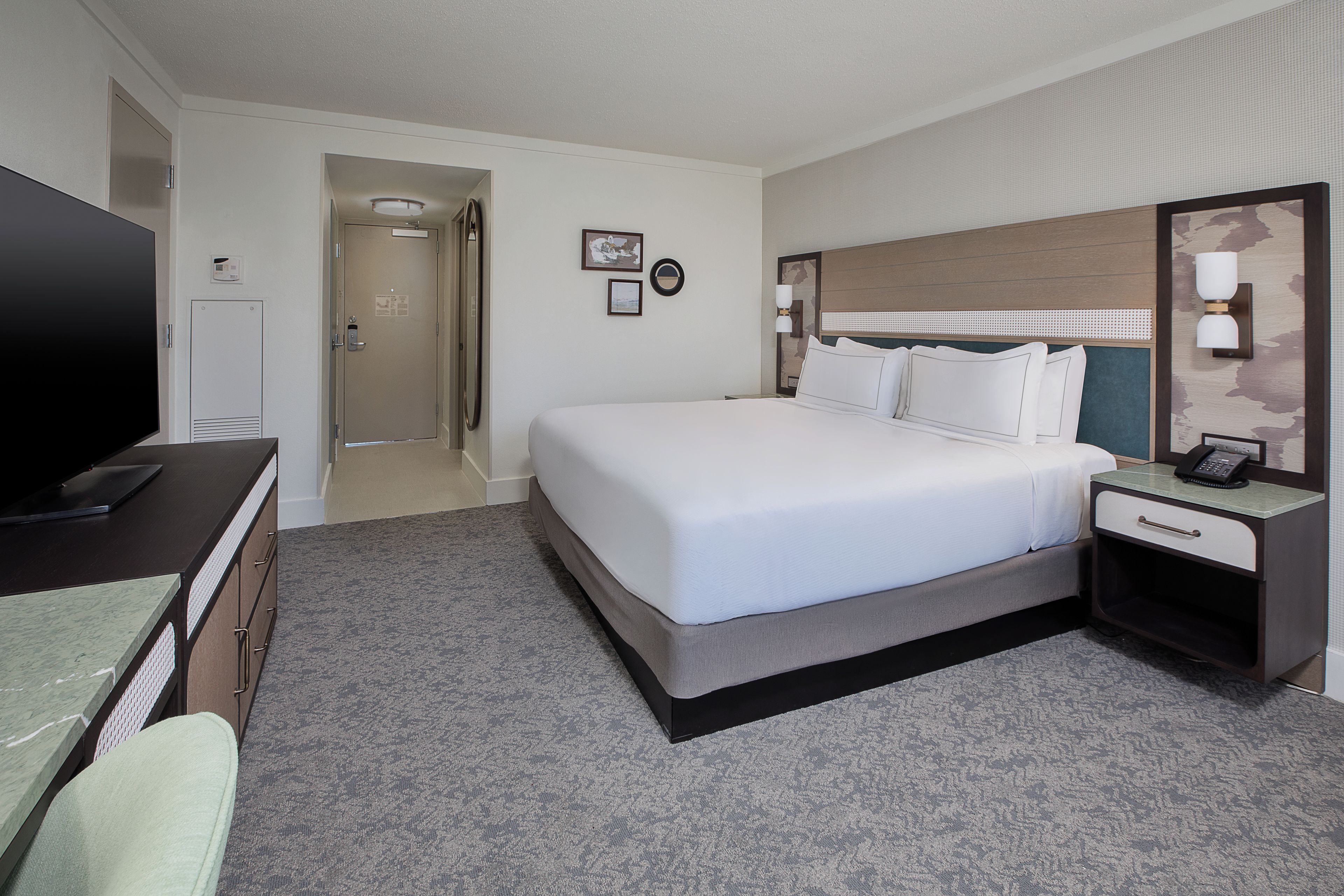 A hotel room with a large bed, a desk with a flat-screen TV, a telephone, and a small bedside table.