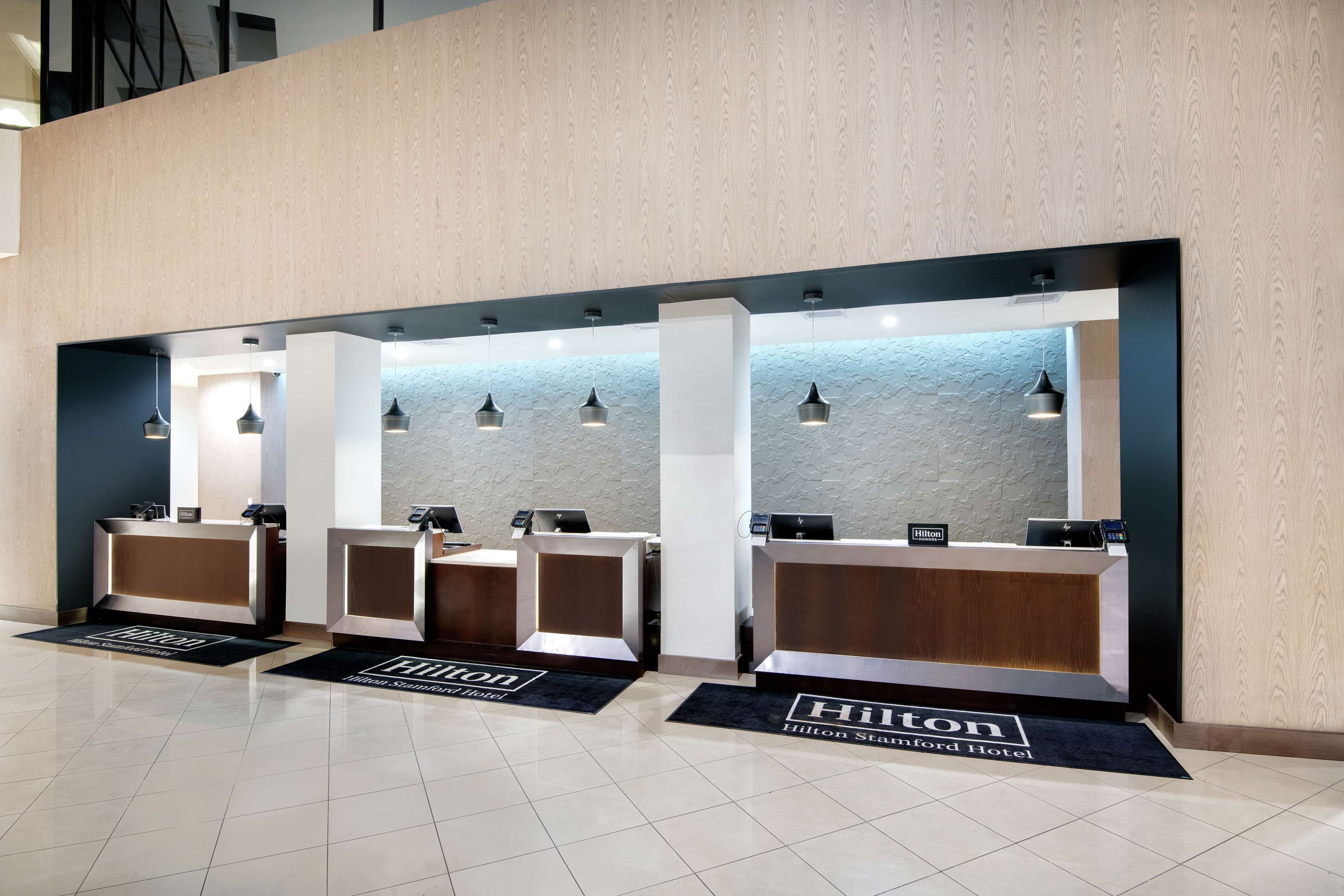 Een hotel receptie met drie balies, elk voorzien van een digitaal scherm en een telefoon. De vloer is belegd met een vloerkleed met het logo van het hotel.
