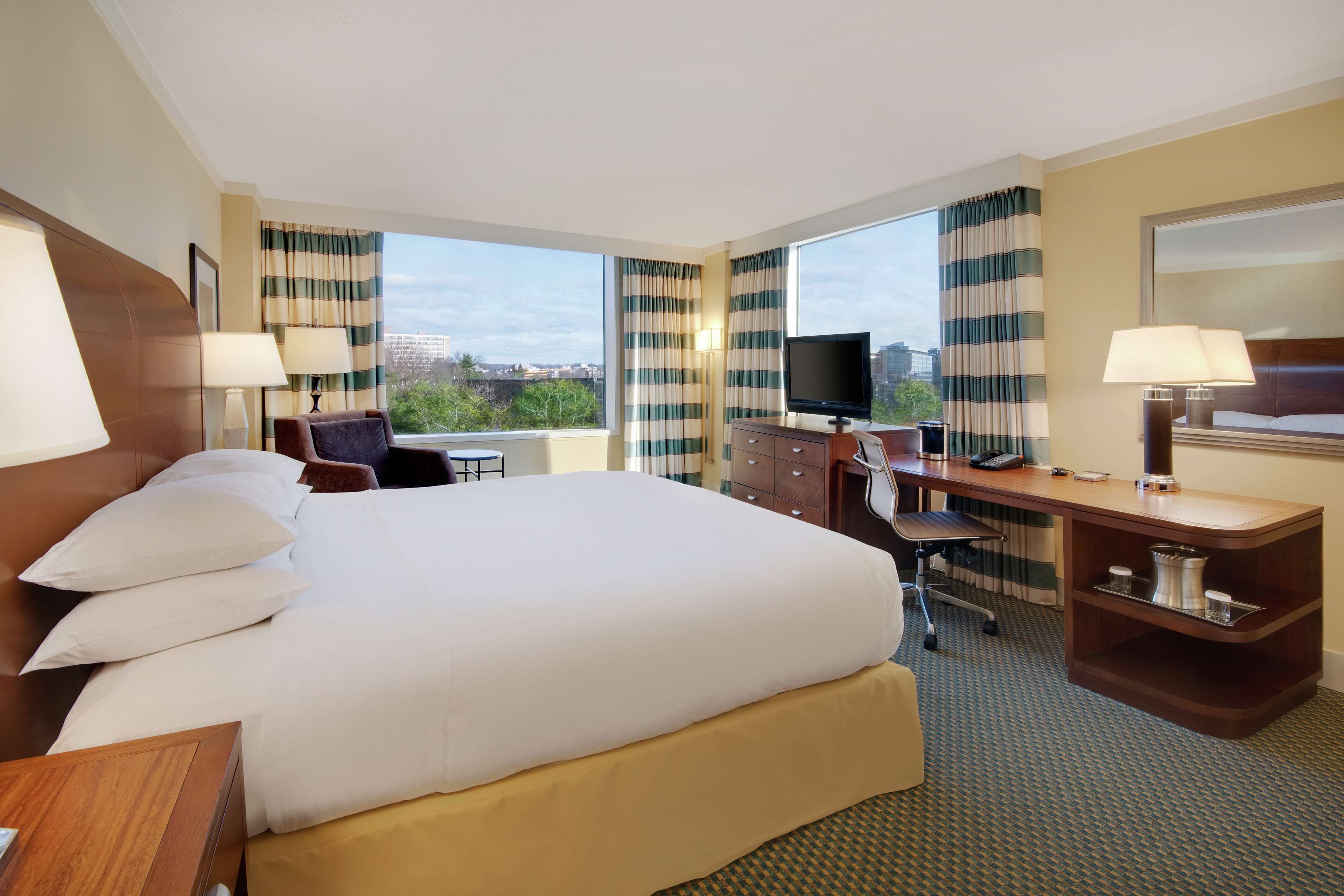 Quarto de hotel com uma cama grande, uma escrivaninha, uma cadeira, televisão e vista para a cidade pelas janelas.