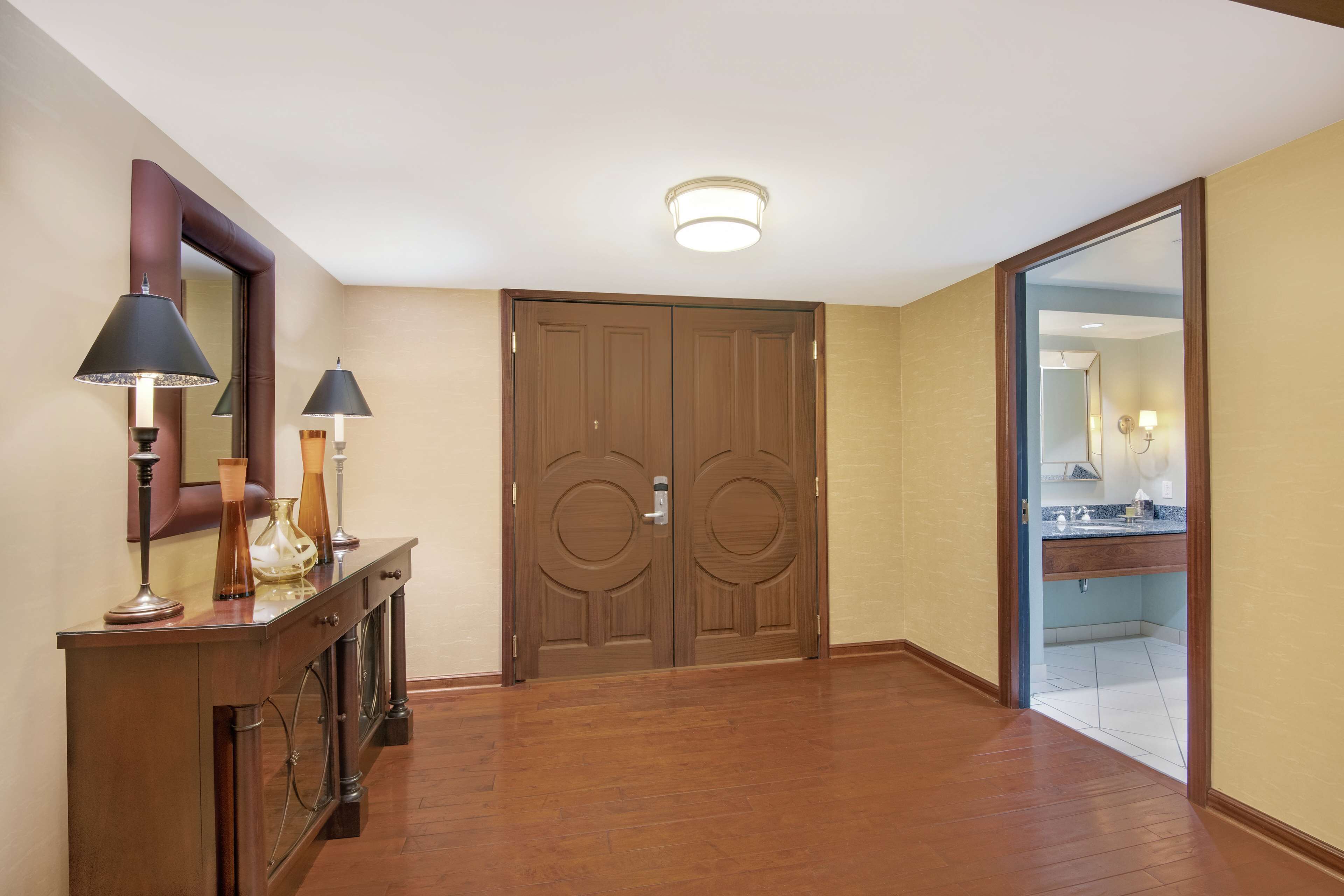 A hallway with wooden flooring, a wooden console table with lamps, and a double door.