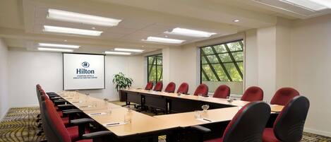 A conference room with a long table, red chairs, and a projector screen displaying the Hilton logo.