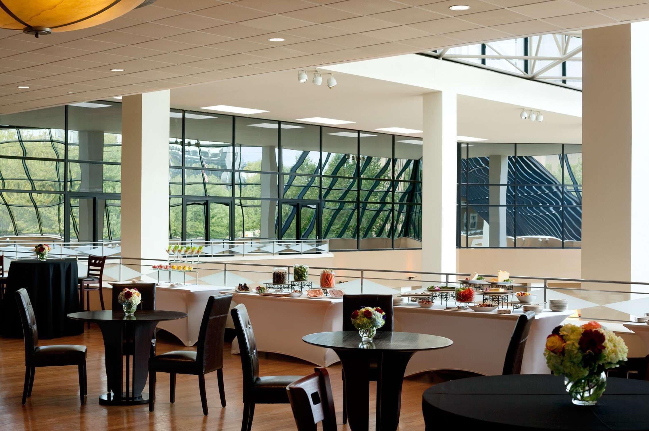 A spacious indoor area with multiple tables and chairs, large windows with a view of greenery, and a modern ceiling design.