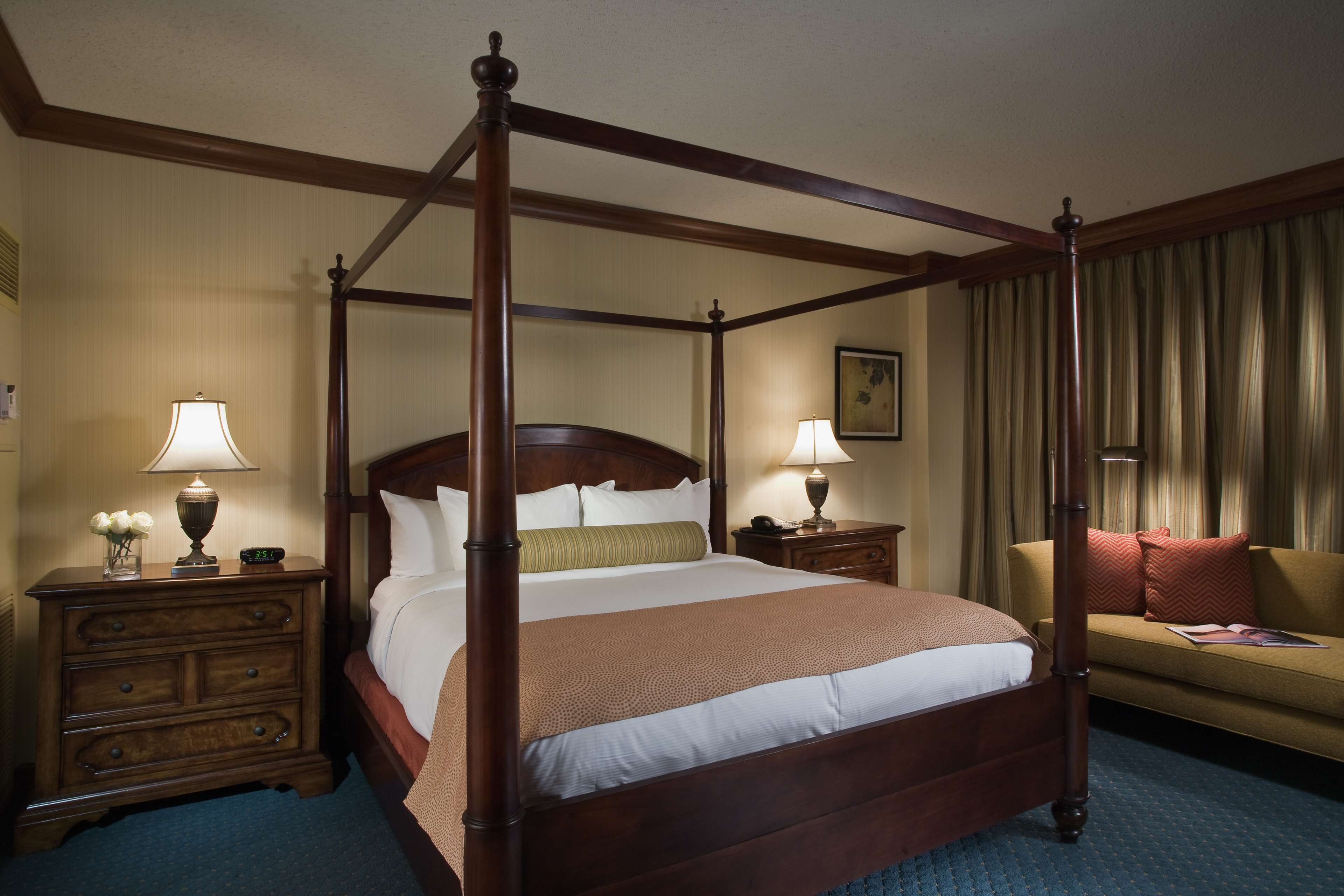 A four-poster bed with a wooden frame, a nightstand with a lamp, a dresser, and a sofa with a cushion.