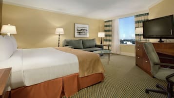 Habitación de hotel con una cama grande, un sofá, un escritorio con televisor y vistas a la ciudad.