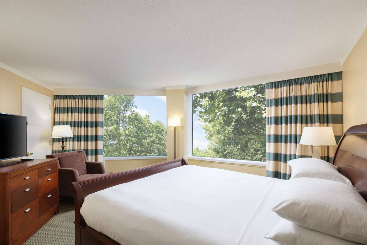 Ein Hotelzimmer mit Bett, Fernseher, Schreibtisch und zwei mit Vorhängen versehenen Fenstern.
