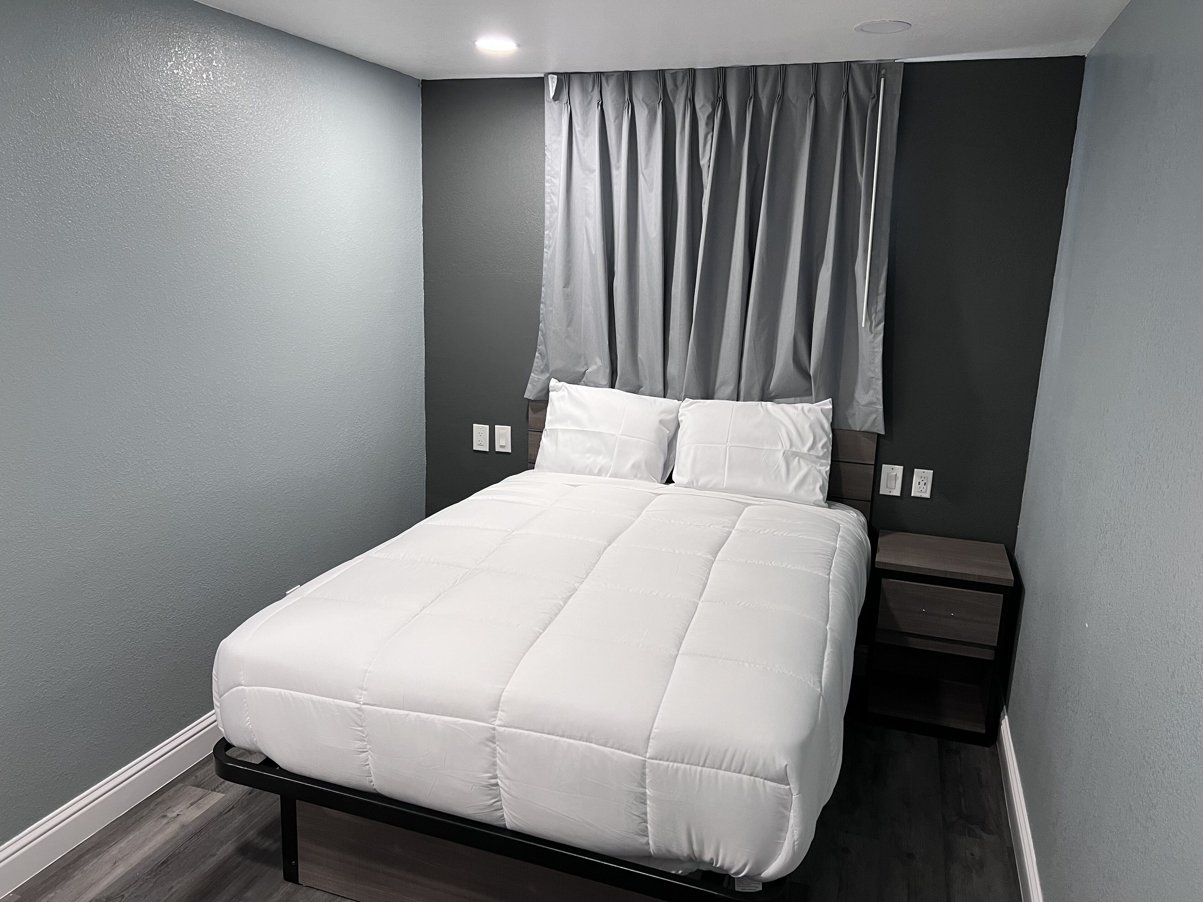 A neatly made bed with white bedding in a room with grey walls and a dark floor.