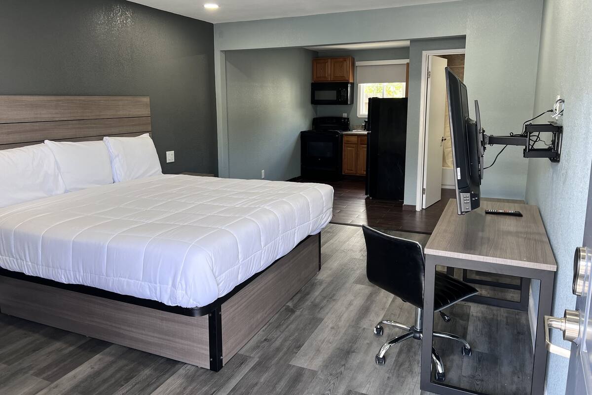 A hotel room with a large bed, a desk with a chair, a TV mounted on the wall, and a kitchenette in the background.