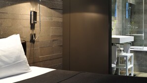 A modern hotel room with a bed, a nightstand, a wall-mounted light, and a glass-enclosed shower area. - Hôtel Sezz Paris (Paris)
