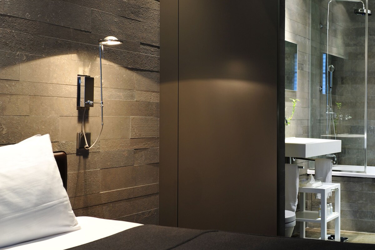 A modern hotel room with a bed, a nightstand, a wall-mounted light, and a glass-enclosed shower area.