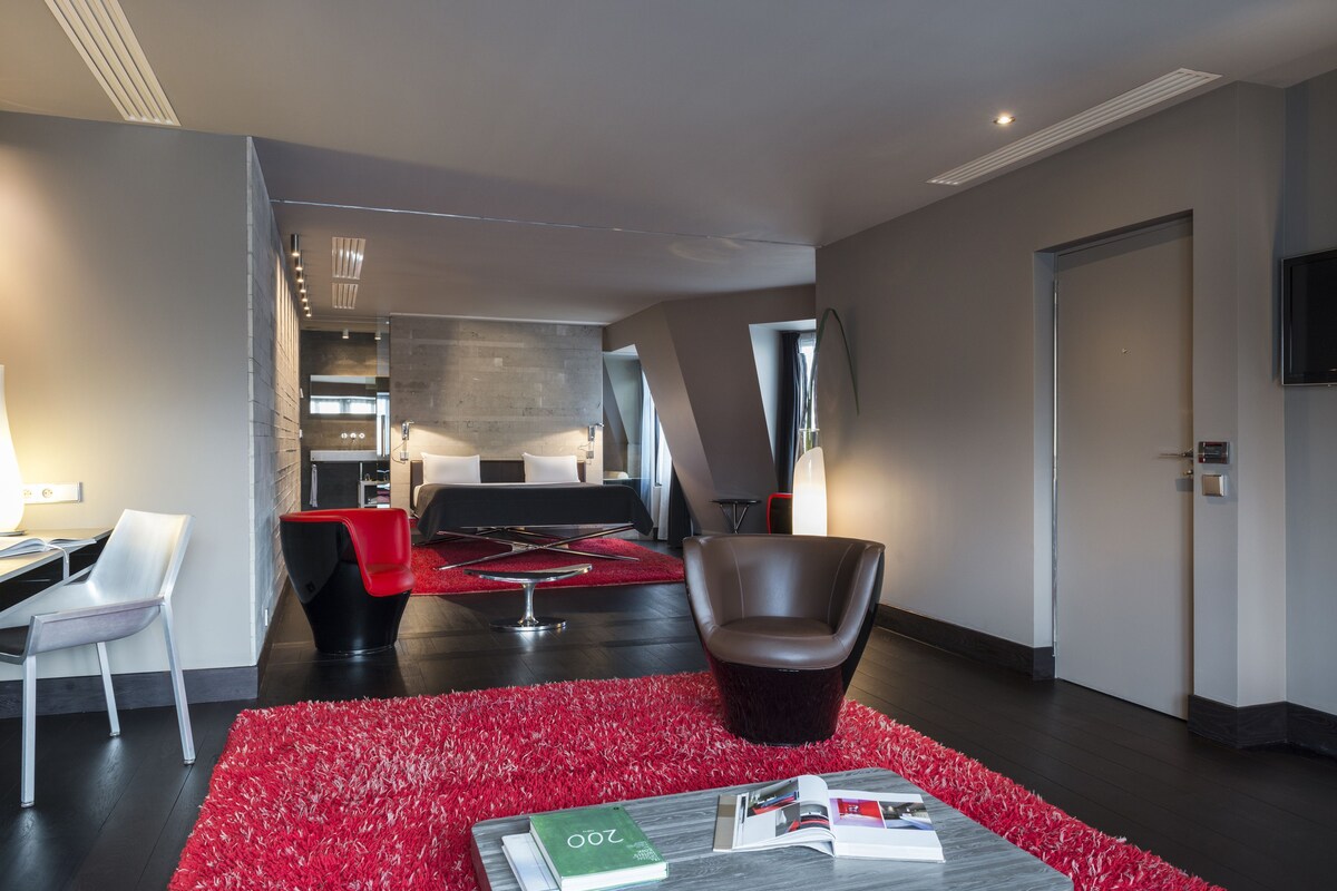 A modern hotel lobby with a red carpet, a dining area with a black table and chairs, a leather armchair, and a flat-screen TV mounted on the wall.