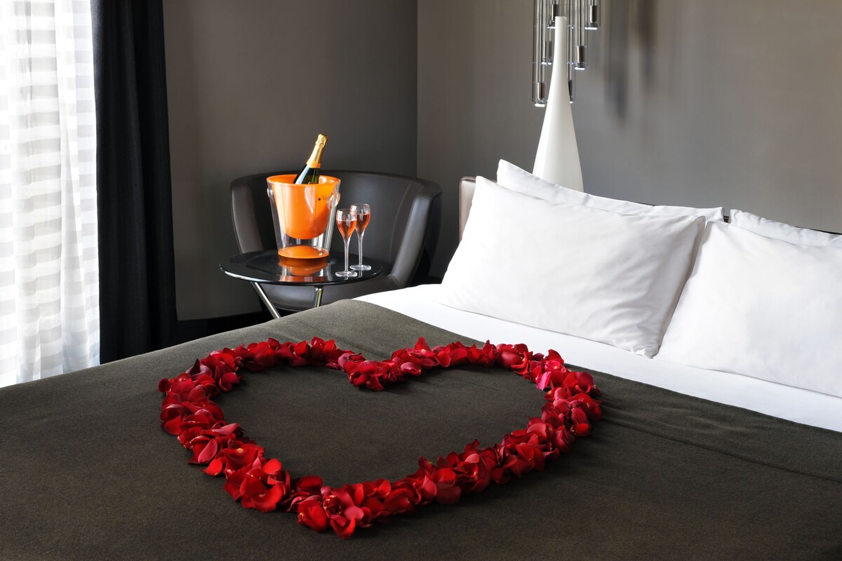 A hotel room with a bed, a heart-shaped decoration made of red petals, a chair, a table with a bottle of champagne, and a white pillow.