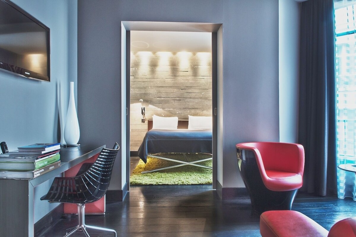 A modern hotel room with a bed, a red chair, a desk with books and a lamp, and a TV on the wall.