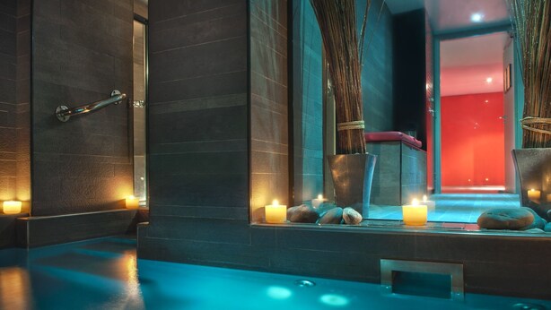 A spa area with a blue-lit floor, a window with a view of a red room, and candles on a ledge. - Hôtel Sezz Paris (Paris)