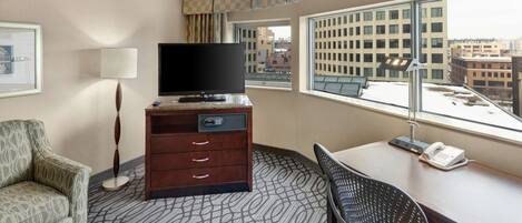 Habitación de hotel con televisión de pantalla plana sobre un mueble de madera, un escritorio con teléfono, un sillón estampado y vistas a la ciudad a través de amplios ventanales.