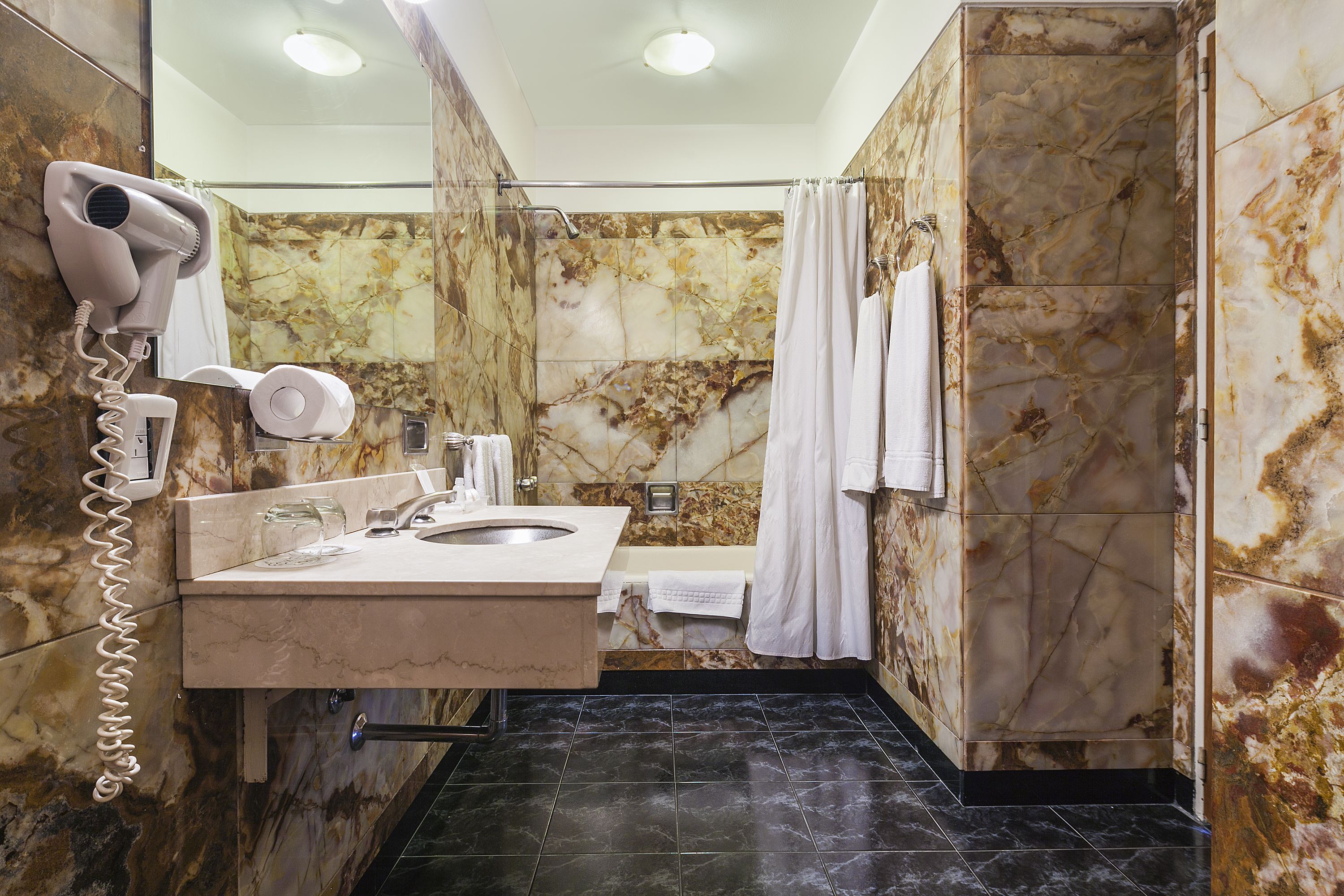 Baño con pared de mármol, lavabo, secador de pelo y cortina de ducha.