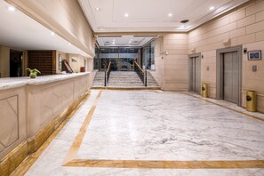 Un vestíbulo de hotel con suelo de mármol, un mostrador de recepción, una escalera y ascensores.