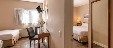 A hotel room with two single beds, a small table with a chair, a desk with a computer, a mirror, and a window with curtains.
