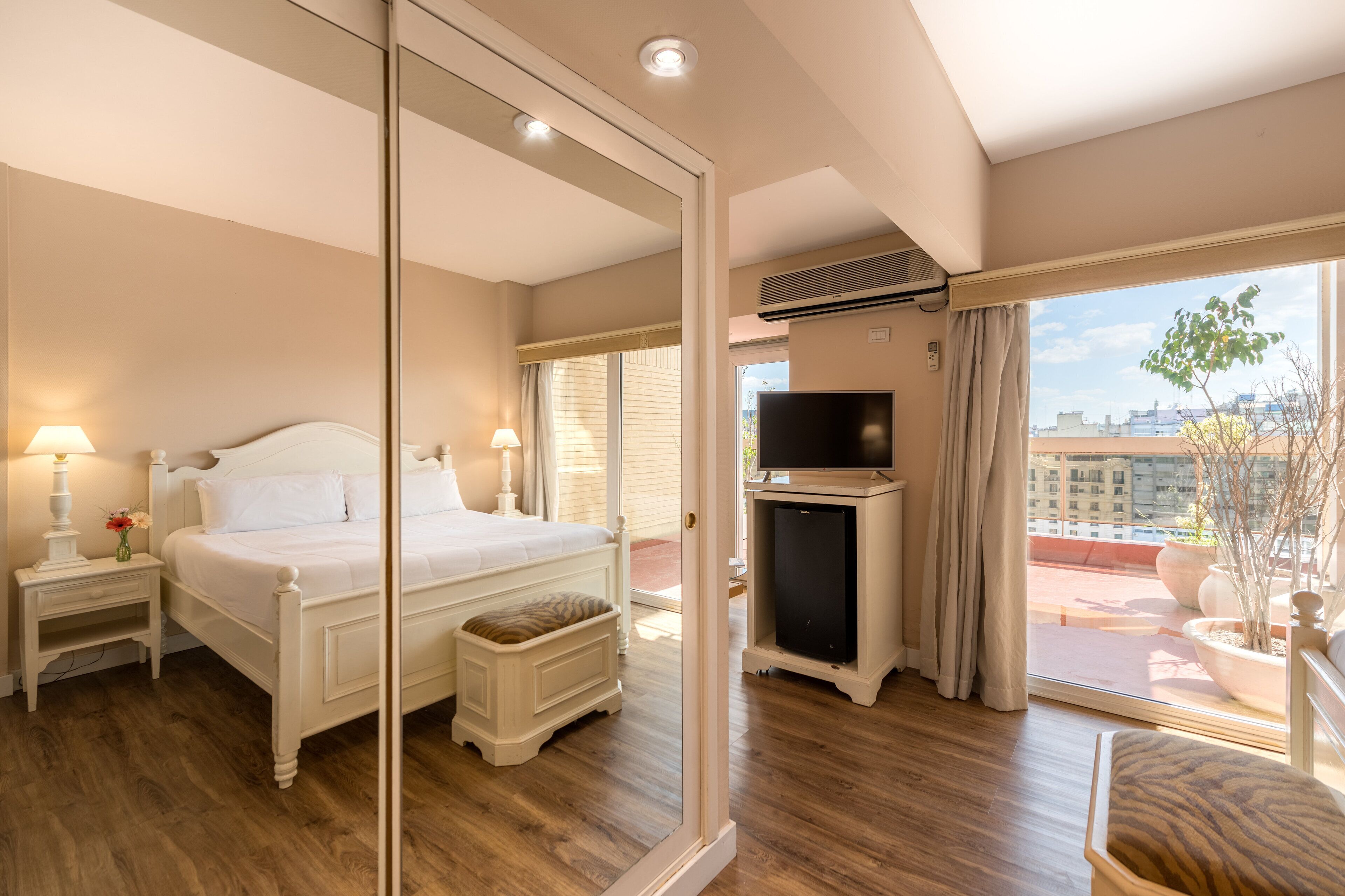 A hotel room with a large bed, bedside tables, a TV, and a balcony with a view of the city.