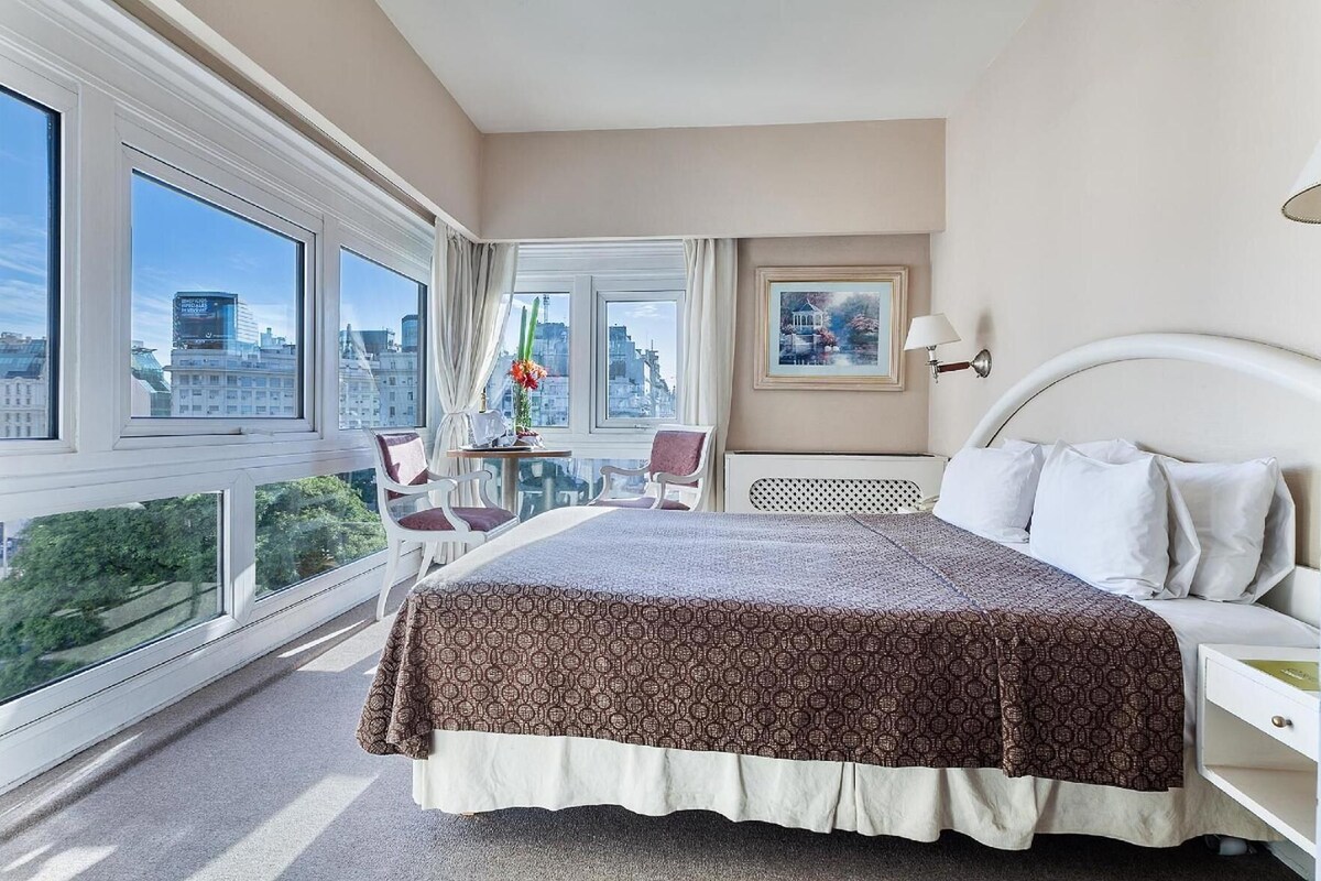 Habitación de hotel con una cama grande, una mesita con sillas, vistas de la ciudad a través de amplias ventanas y un cuadro de paisaje urbano en la pared.