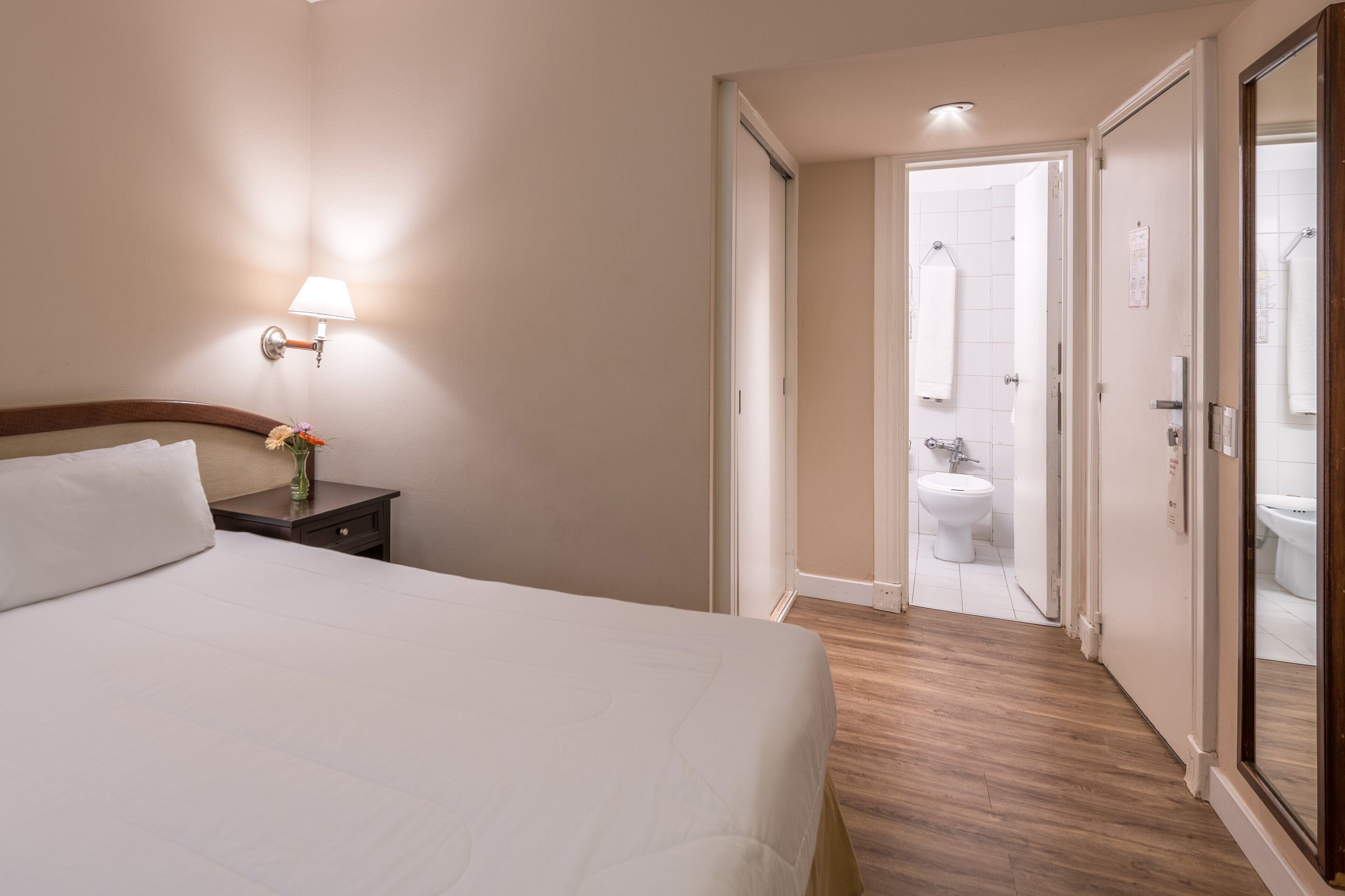 A hotel room with a bed, a nightstand with a lamp, and a view of the bathroom through an open door.
