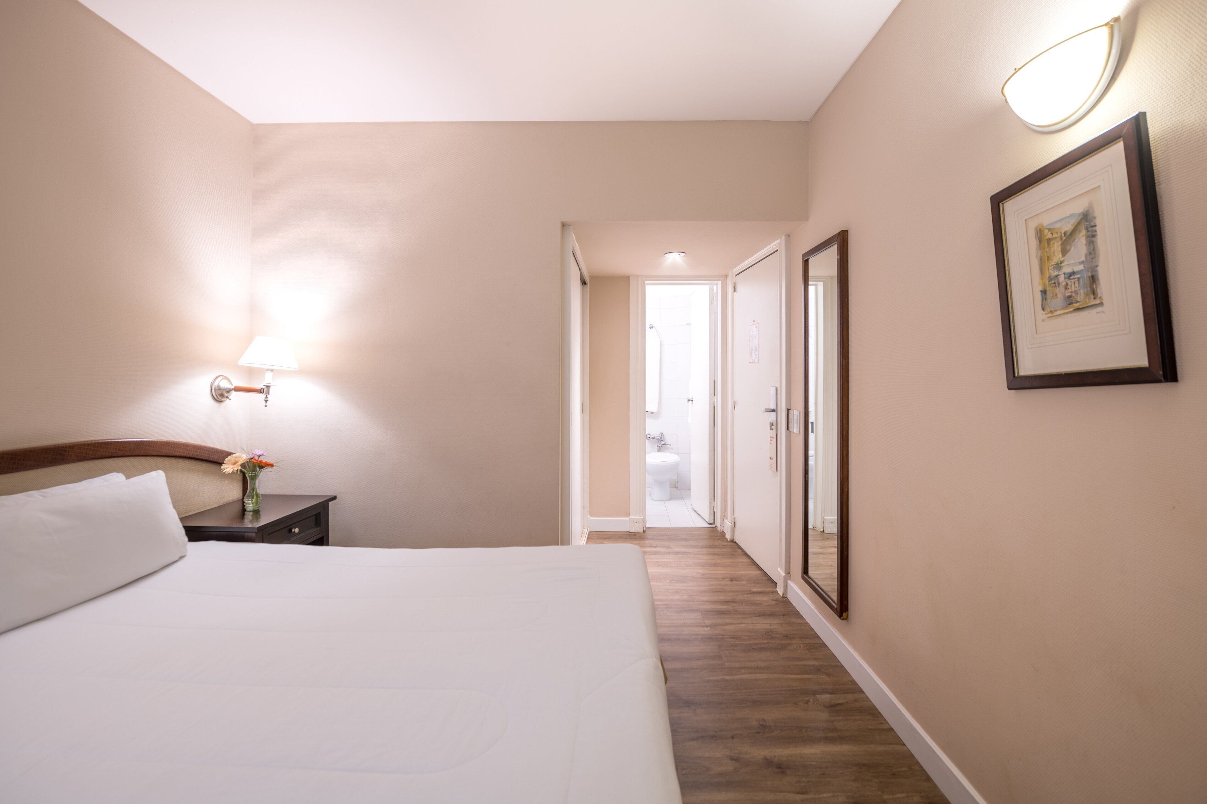 Habitación de hotel con cama, mesita de noche, lámpara, cuadro enmarcado y un pasillo que conduce a un baño.