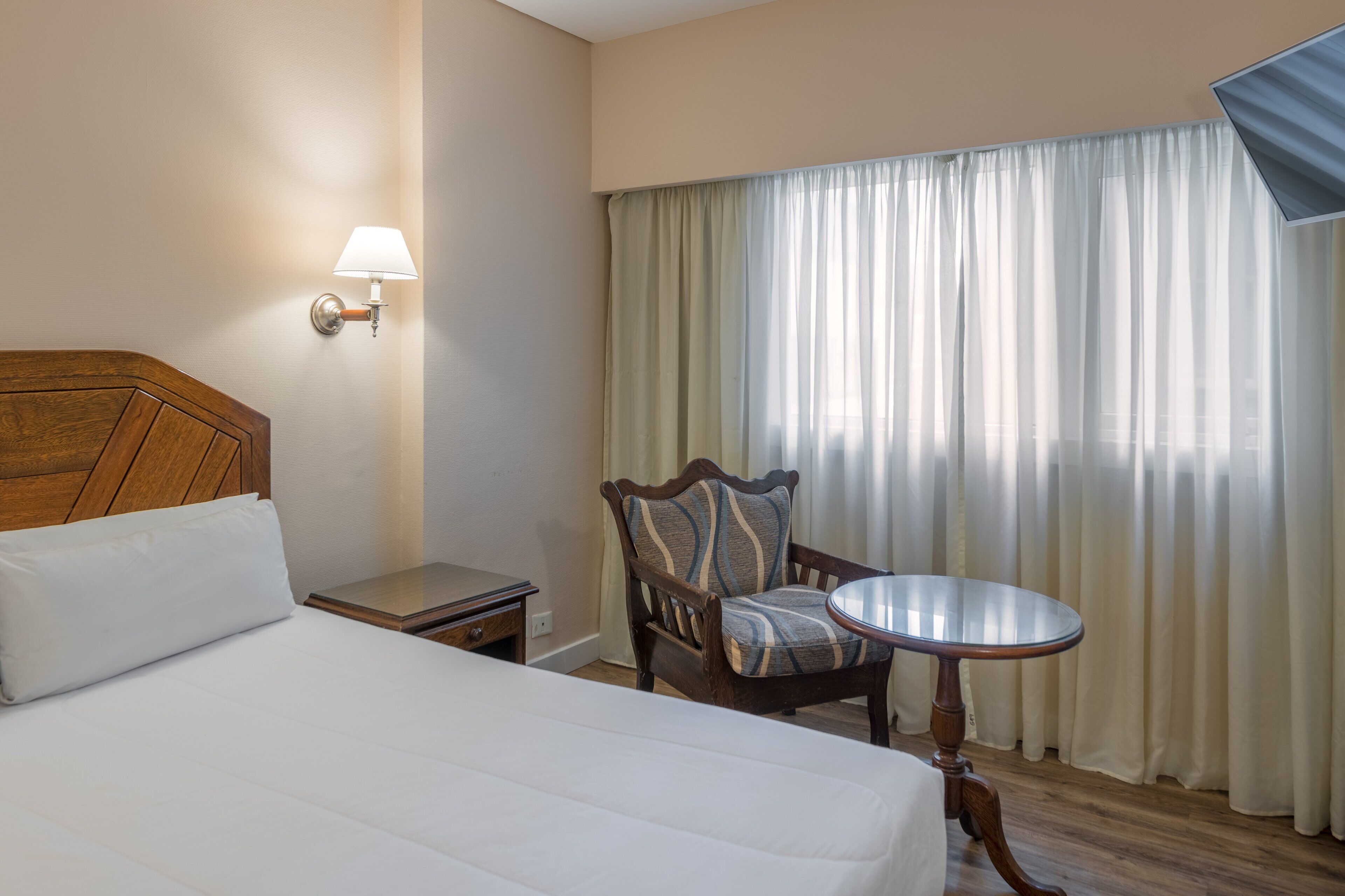 Una habitación de hotel con cama, una silla, una mesita, una lámpara y una ventana con cortinas.