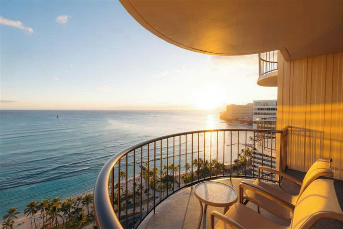Ein Balkon mit gebogener Geländer, der auf einen Strand und das Meer blickt. Es gibt einen runden Tisch und zwei Stühle.