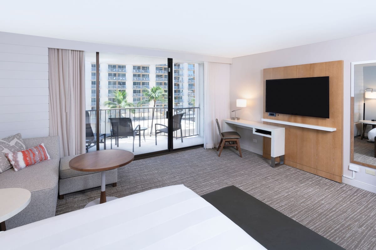 Ein modernes Hotelzimmer mit einer Sitzecke, einem kleinen Tisch, einem Schreibtisch, einem Stuhl, einem an der Wand befestigten Fernseher und einem Balkon mit Blick auf Palmen und Gebäude.