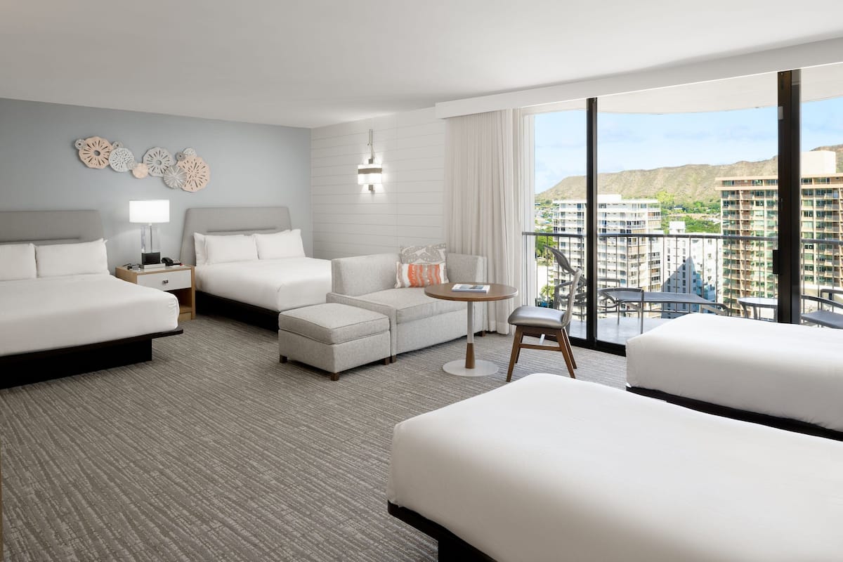 Ein Hotelzimmer mit einem großen Bett, einem Schlafsofa, einem kleinen Tisch und einem Balkon mit Blick auf Gebäude.