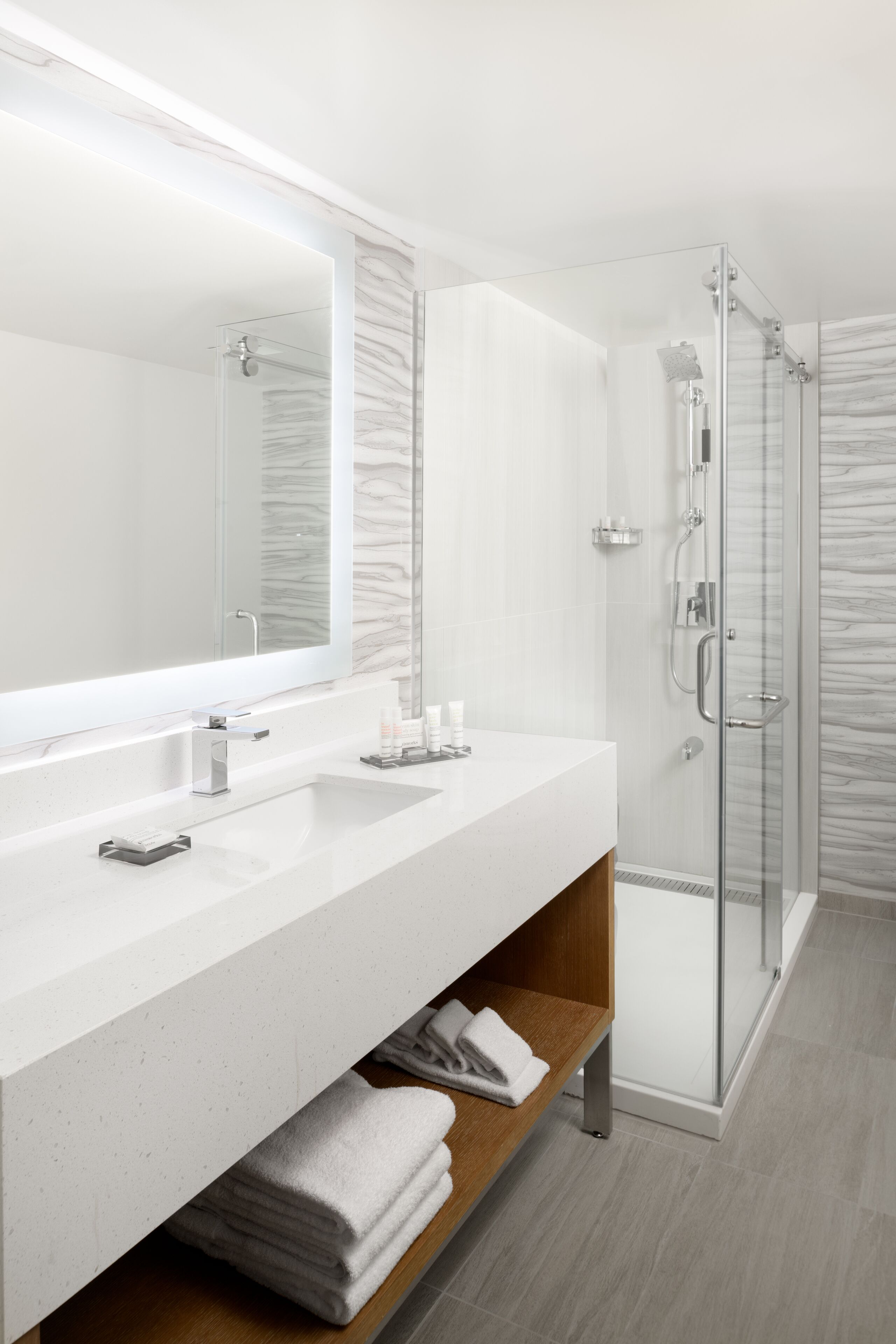 Un baño moderno con un espejo grande, dos lavamanos, una cabina de ducha de vidrio y un estante de madera con toallas.