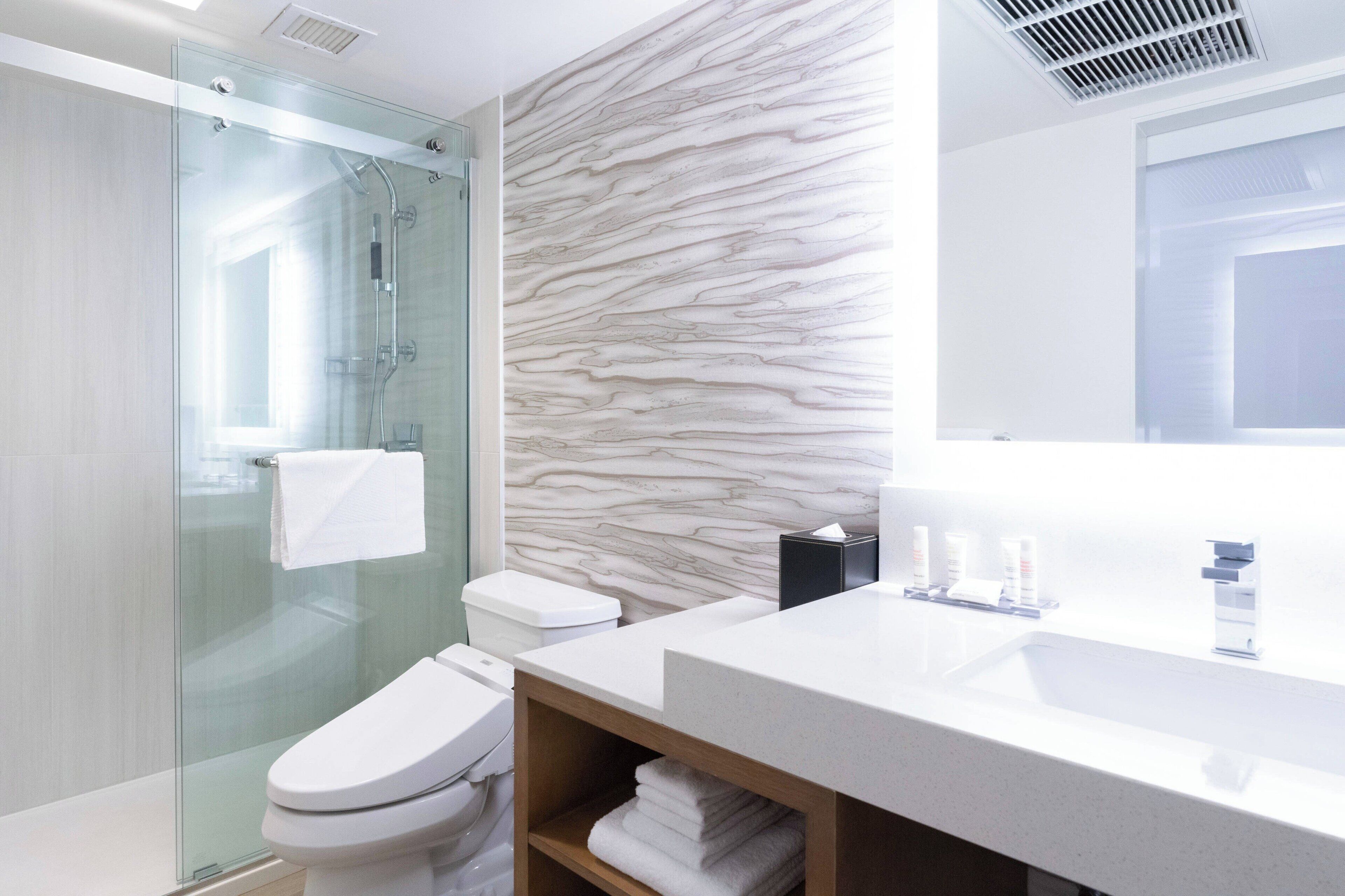 A modern bathroom with a glass-enclosed shower, a marble-patterned wall, a white sink, and a mirror.