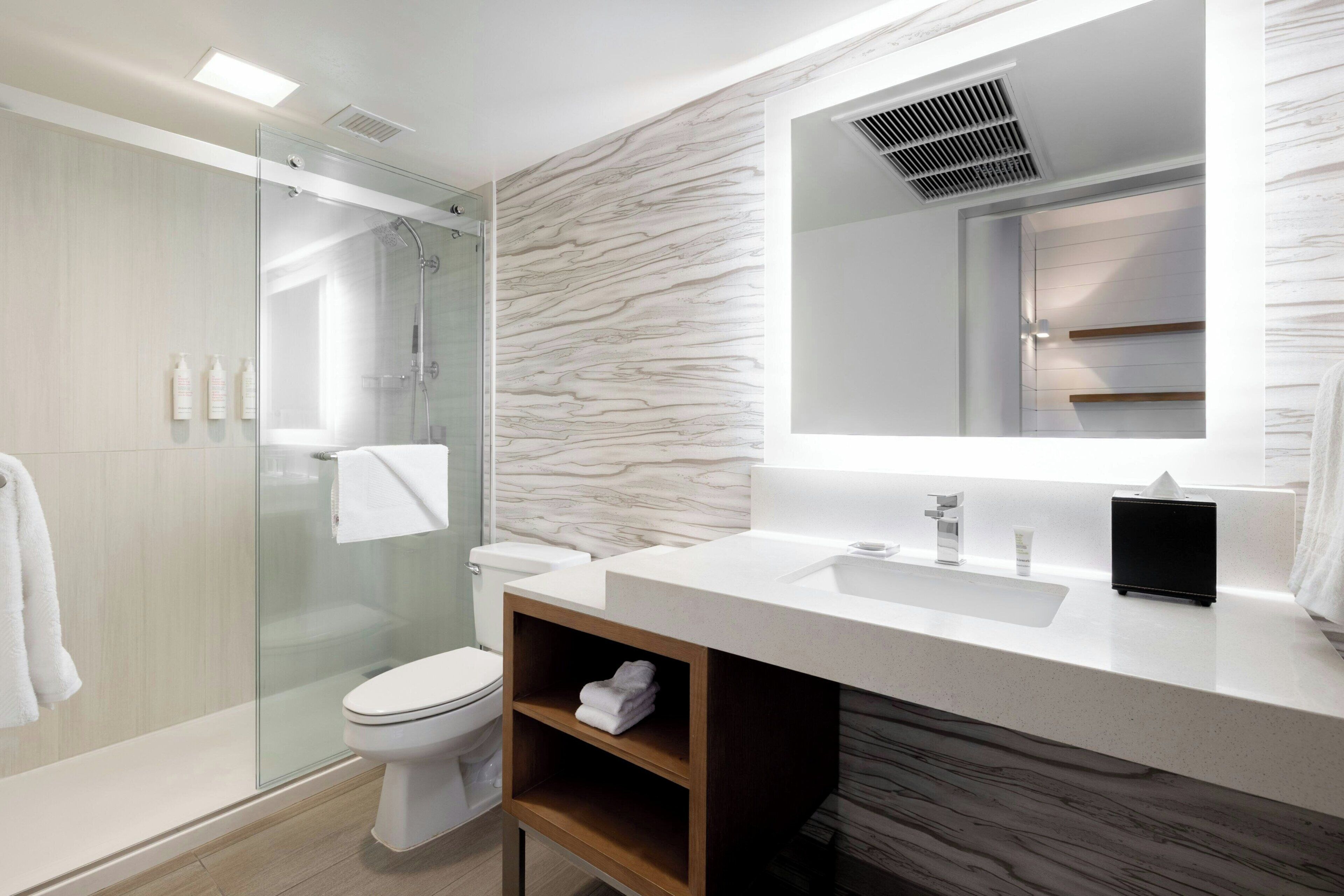 A modern bathroom with a glass shower enclosure, a white toilet, a marble-patterned wall, a white sink, and a black radio.