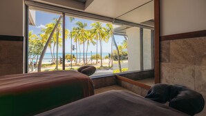 Quarto de hotel com uma grande janela com vista para uma praia com palmeiras e o oceano.