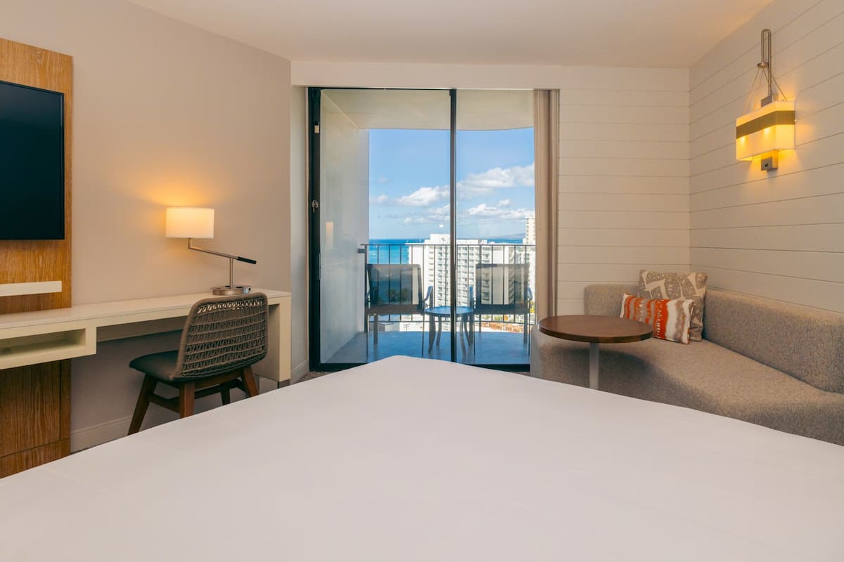 Ein Hotelzimmer mit einem großen Bett, einem Schreibtisch mit Stuhl, einem kleinen Tisch, einer Couch mit Kissen, einem an der Wand montierten Fernseher und einem Balkon mit Blick auf Stadt und Meer.