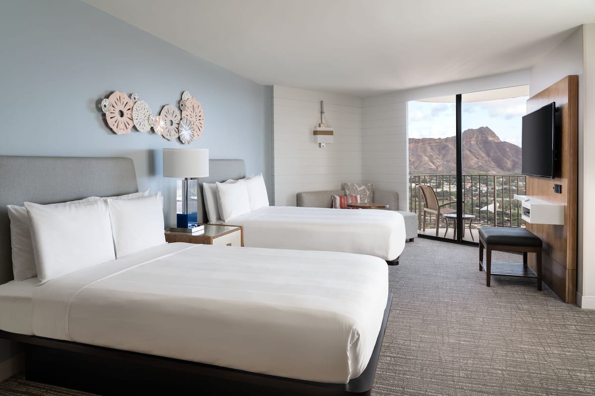 Ein Hotelzimmer mit zwei Betten, einem Fernseher, einem Balkon mit Bergblick und einem Schreibtisch mit Stuhl.