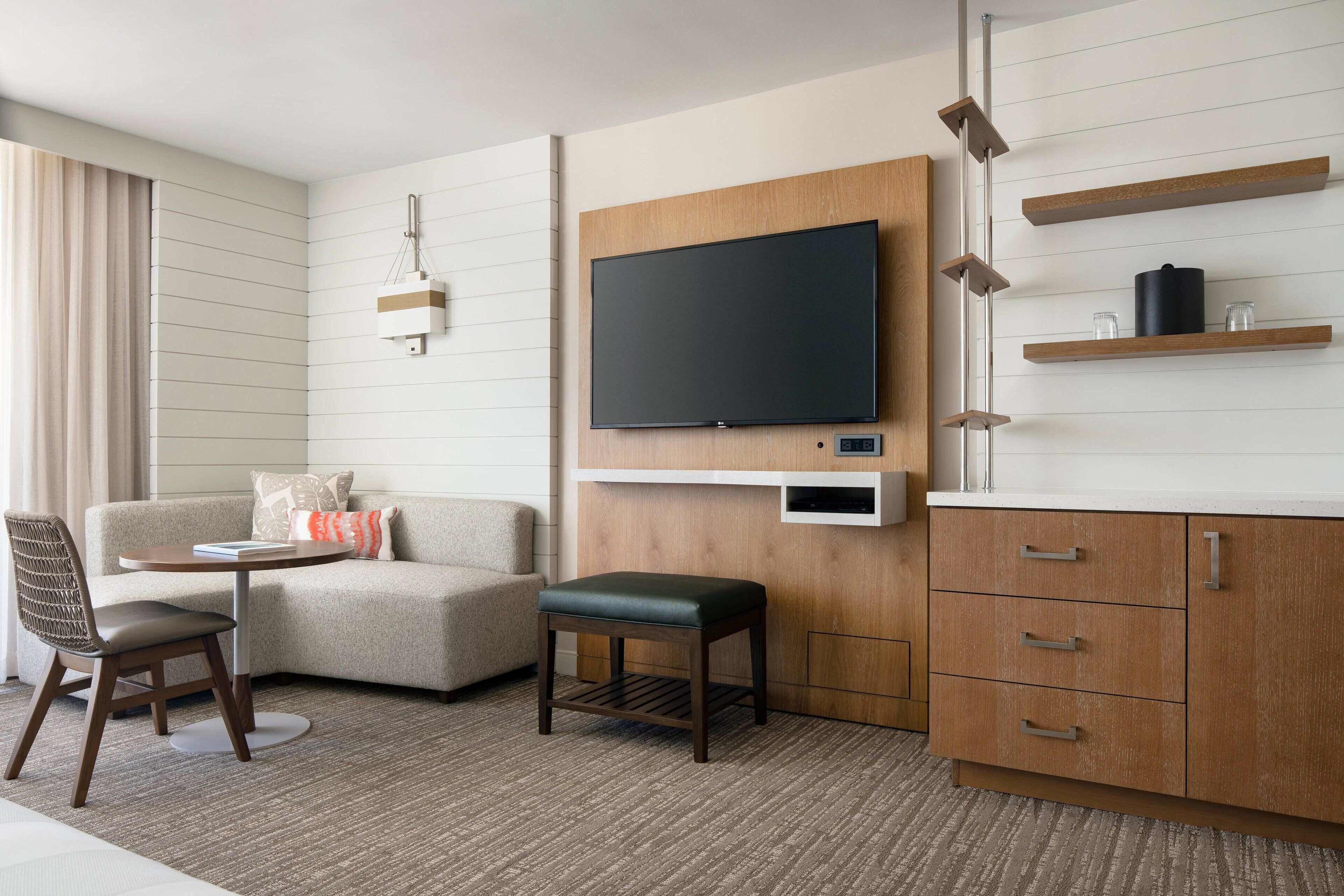 A modern hotel room with a flat-screen TV mounted on a wooden panel, a sofa, a small table, a chair, and a cabinet with drawers.
