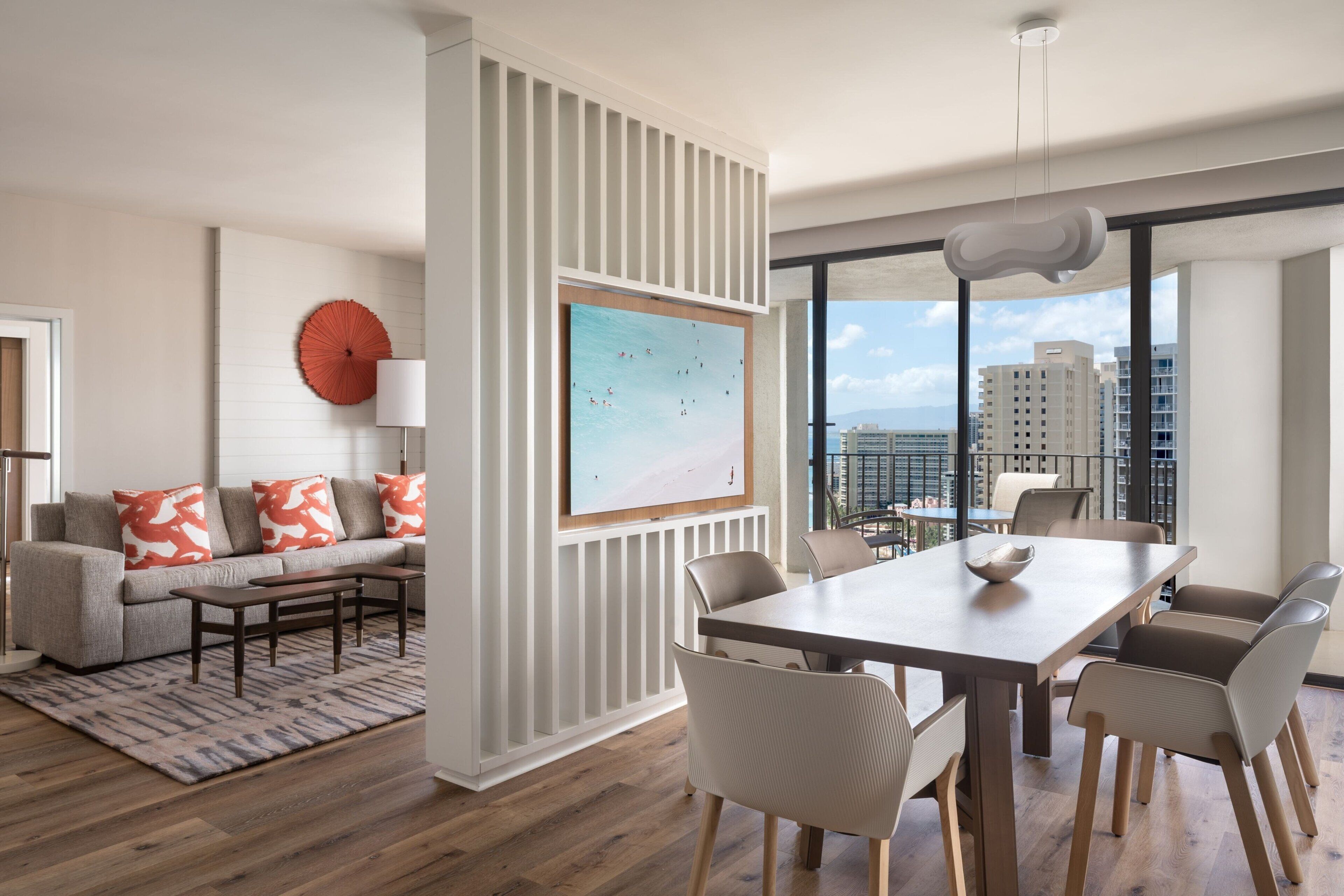 Ein modernes Wohnzimmer mit Sofa, Couchtisch und Essbereich mit Stühlen. Ein großes Fenster bietet Blick auf Gebäude und die Stadtlandschaft.