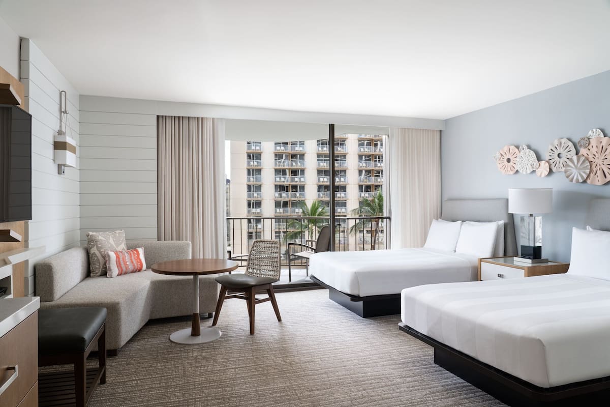 Ein Hotelzimmer mit zwei Betten, einem Sofa, einem kleinen Tisch und einem Sessel. Es gibt einen Balkon mit Blick auf ein Hochhaus.