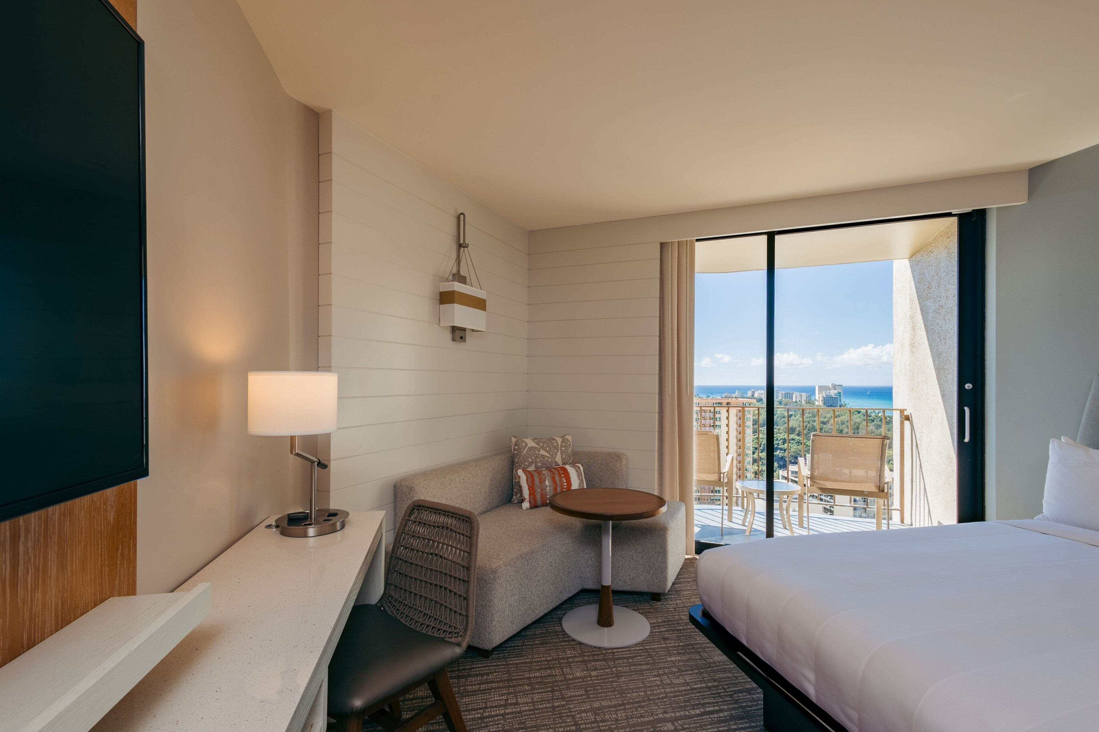 A hotel room with a bed, a desk, a chair, a small table, and a balcony with a view of the city and ocean.