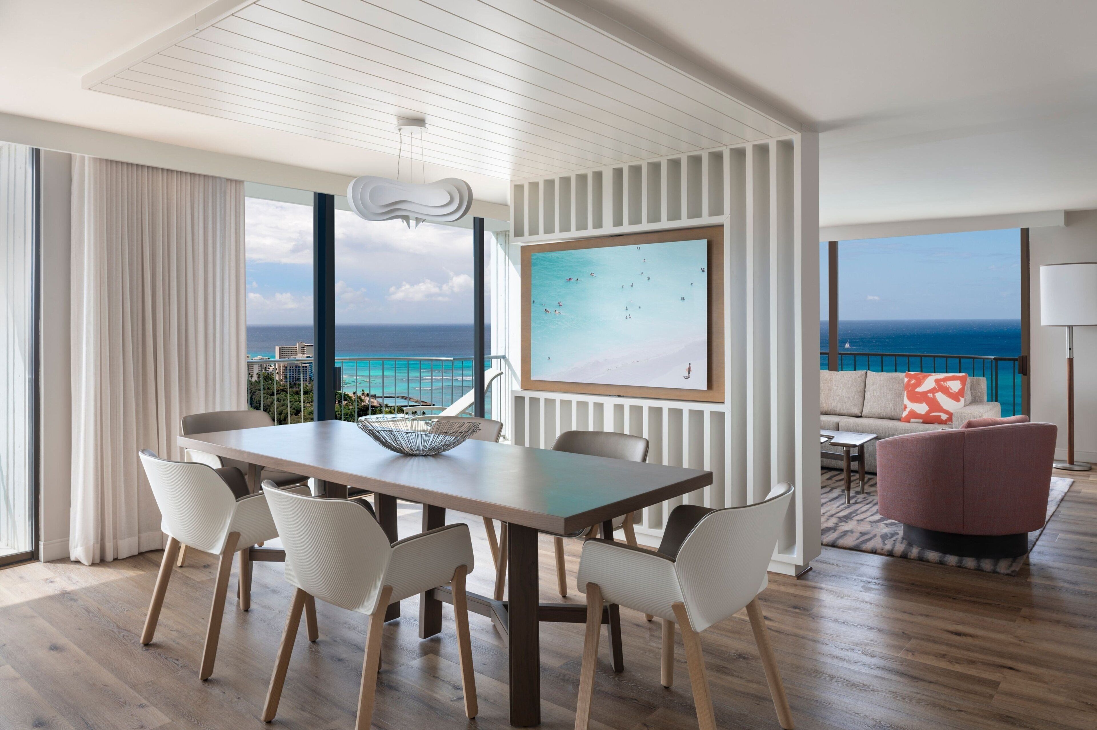 A modern hotel room with a dining area, a large window with a view of the ocean, a painting on the wall, and a cozy seating area with a view.
