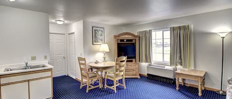 Una habitación con alfombra azul, un lavamanos, una mesa con sillas, una lámpara, una ventana con cortinas y una pequeña mesa de madera.