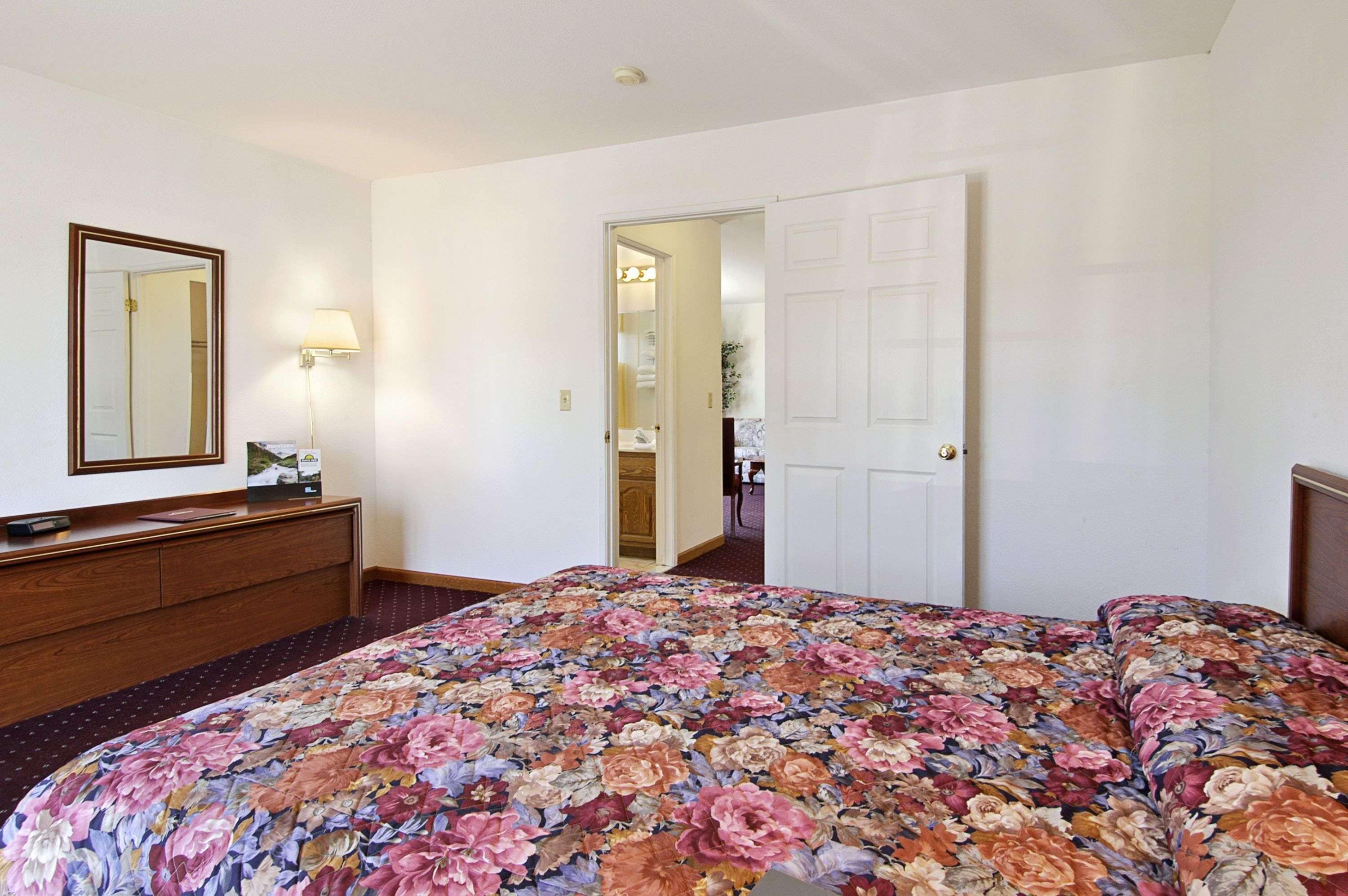 A hotel room with a floral bedspread, a wooden dresser, a mirror, and a door leading to a bathroom.