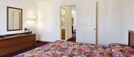 A hotel room with a floral bedspread, a wooden dresser, a mirror, and a door leading to a bathroom.