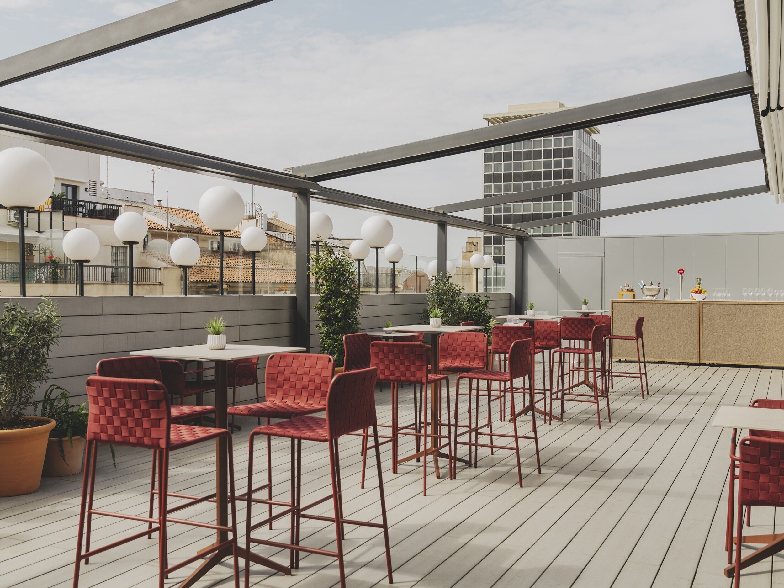 A rooftop bar with red chairs and tables, potted plants, and a bar counter with glasses and a fruit bowl.