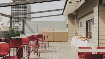 Un espacio de comedor al aire libre con sillas y mesas rojas, una barra de buffet y vista a un edificio alto al fondo.