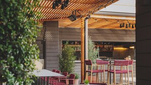 Un espacio al aire libre con mesas y sillas, una barra con estantes llenos de botellas, y una pérgola de madera sobre la cabeza.