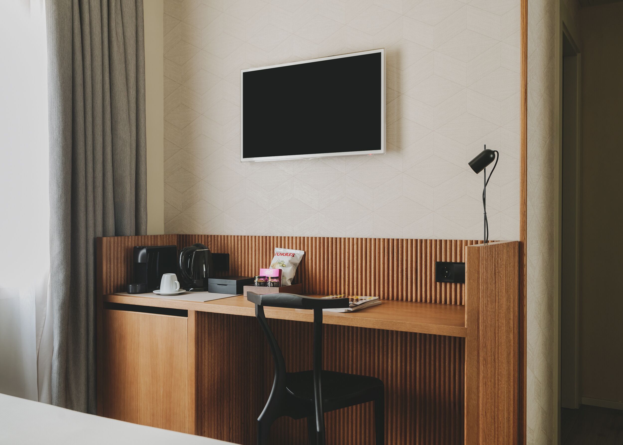 A hotel room with a wooden desk, a black chair, a coffee maker, a TV mounted on the wall, and a bag of coffee on the desk.