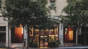 Entrada de hotel con fachada de vidrio, amplias ventanas y una alfombra roja que conduce a la puerta.