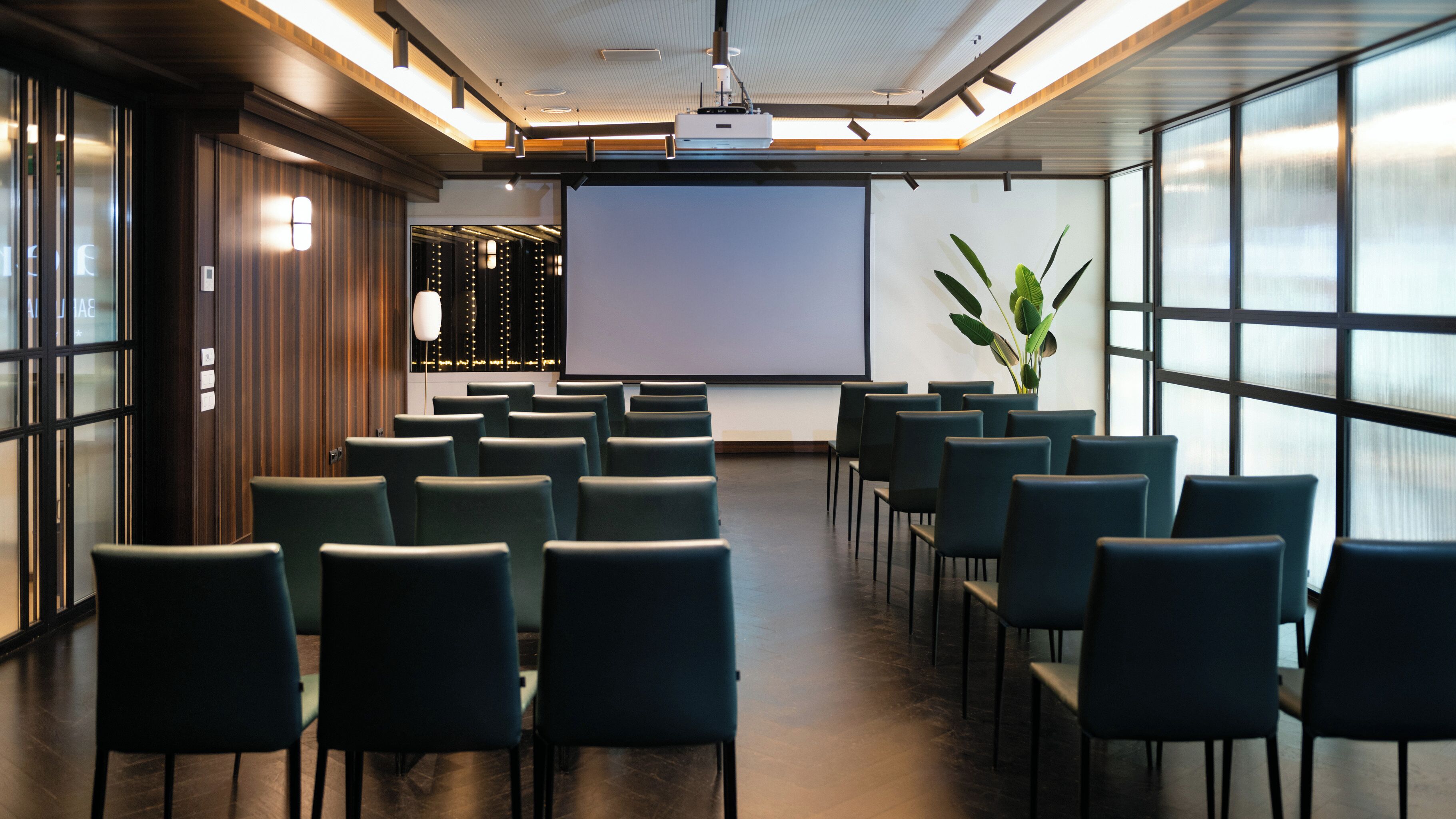 Una sala de conferencias con filas de sillas orientadas hacia una pantalla de proyección, paneles de madera a la izquierda y un ventanal grande a la derecha.